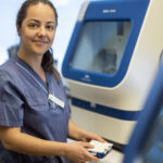 Kvinna i sjukhuskläder står framför instrument på laboratoriet.