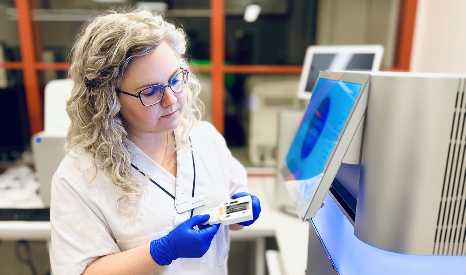Kvinna i vita sjukhuskläder jobbar framför analysinstrument.