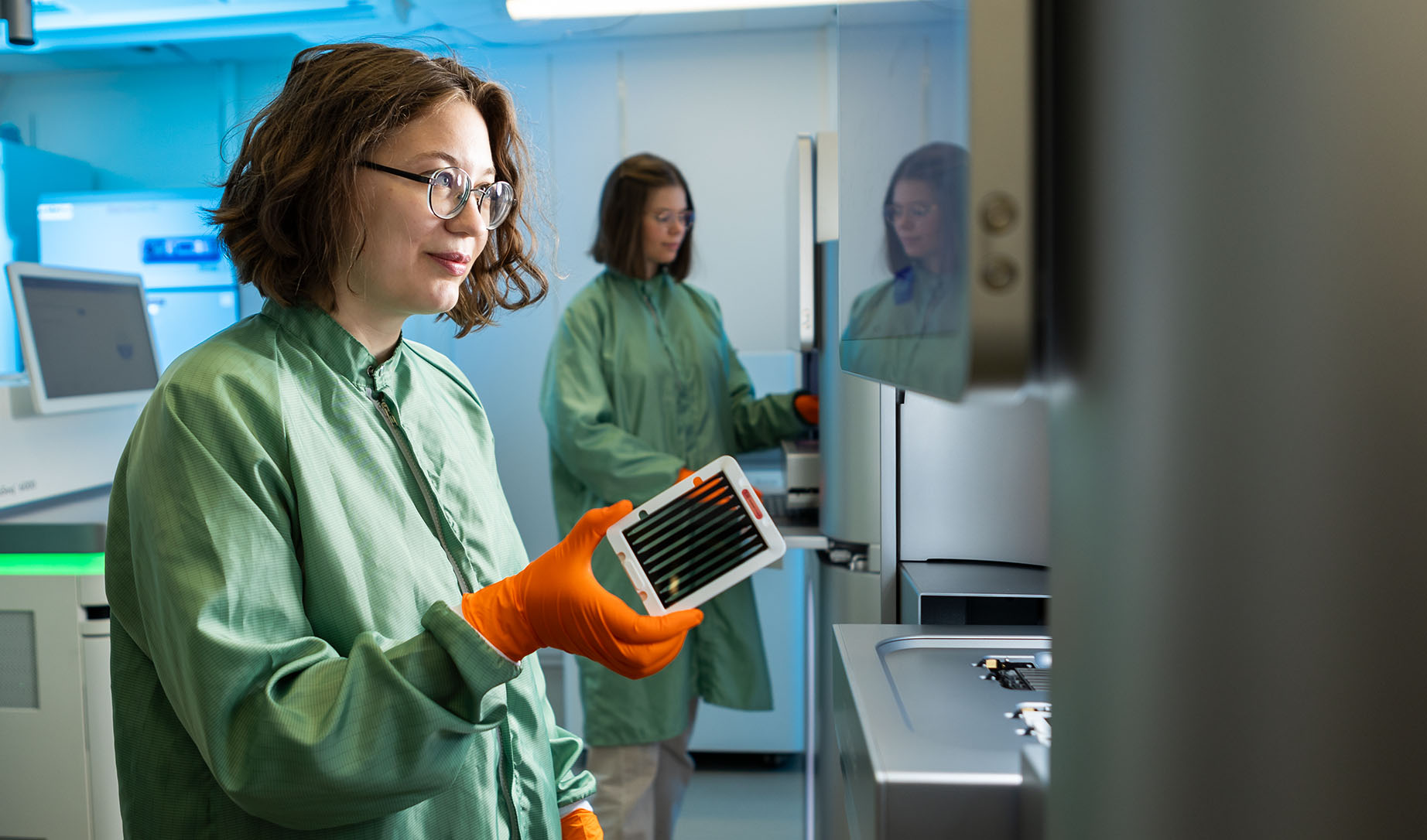 Två kvinnor arbetar iförda grön labrock vid instrument för sekvensering av DNA.